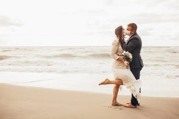 Groom in a chic suit and a beautiful bride in a wedding gown walking along the beach. Newlyweds kisses in protective medical masks. Toned.