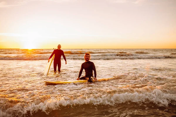 Two young male surfers in black wetsuits with longboards in a water at sunset ocean. Water sport adventure camp and extreme swim on summer vacation.