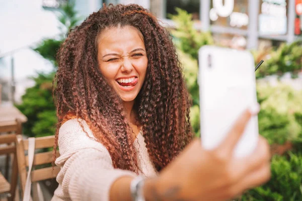 Young smiling curly woman takes a selfie with white smartphone sitting at the cafe outside and shows her tongue.
