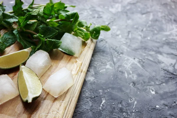 Kokteyl mojito için malzemeler. Çatlak ve çizikler ile gri beton arka plan üzerinde ahşap kesme tahtası üzerinde taze kireç parçaları, nane ve buz
