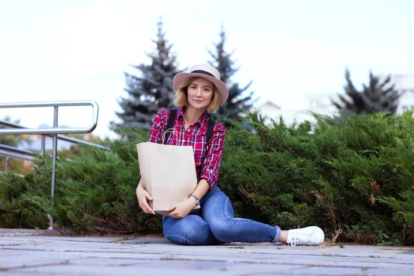 Şık kız zanaat kağıt paketi tutan. Şehir parkında şapkalı bir kız dinleniyor. Elinde çevre dostu alışveriş çantası. Sıfır atık yaşam tarzı. Plastik özgür yaşam. Plastik yok. Paket tasarım şablonu mock up