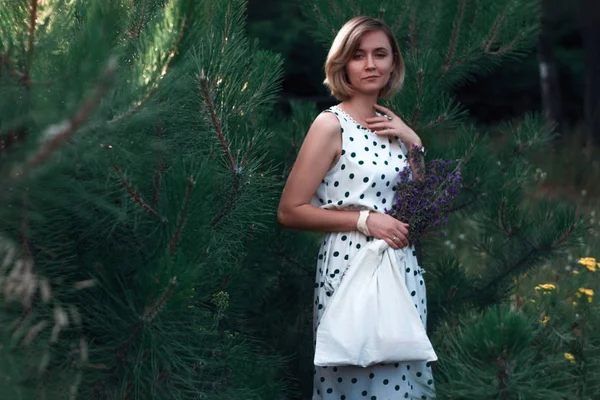 Beautiful woman holding cotton Eco bag with field flowers in her hands. Reusable cotton bag in the hands. Girl in the forest in white dress in peas. Zero waste and conscious lifestyle