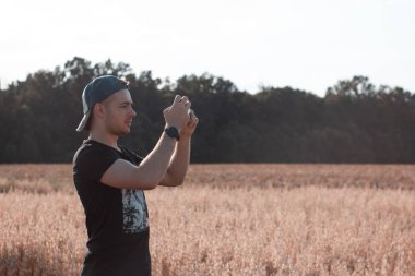 Genç adam akıllı telefonuyla fotoğraf çekiyor. Mobil fotoğrafçı görsel içerik oluşturuyor. Alanın ve doğanın fotoğrafını çekiyoruz. Açık havada, ofis dışında, doğa geçmişi olan bir adam..