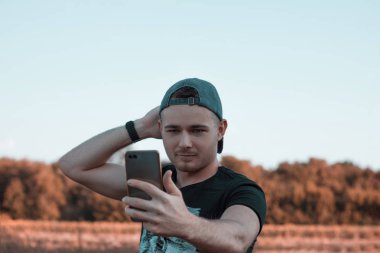 Çekici bir gencin portresi. Açık havada bir şapkayı düzeltirken ve ayakta dururken cep telefonuyla selfie çekiyor. Günlük yaşamda aletleri kullanmak.