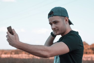 Çekici bir gencin portresi. Açık havada bir şapkayı düzeltirken ve ayakta dururken cep telefonuyla selfie çekiyor. Günlük yaşamda aletleri kullanmak.