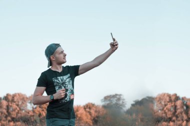 Çekici bir gencin portresi cep telefonuyla, açık havada selfie çekiyor. Günlük yaşamda aletleri kullanmak.
