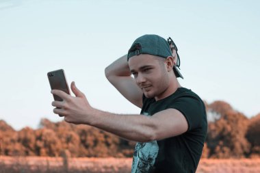 Çekici bir gencin portresi. Açık havada bir şapkayı düzeltirken ve ayakta dururken cep telefonuyla selfie çekiyor. Günlük yaşamda aletleri kullanmak.