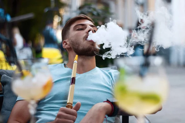Yakışıklı adam nargile dumanı üflemiş. Genç adam dışarıda güzel kokulu bir nargile içiyor. Yaz bölgesinde nargile ve kokteyl ile dinlen..