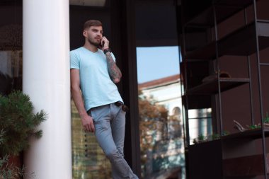 Yakışıklı adam dışarıda cep telefonuyla konuşuyor. Kolunda dövmesi ve akıllı saati olan modern stil sahibi genç adam telefona cevap veriyor. Akıllı telefonu kırmızı bir çantada.. 