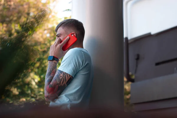 Yakışıklı adam dışarıda cep telefonuyla konuşuyor. Kolunda dövmesi ve akıllı saati olan modern stil sahibi genç adam telefona cevap veriyor. Akıllı telefonu kırmızı bir çantada. Arka plan