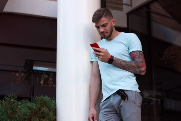 Yakışıklı adam dışarıdaki cep telefonundan mesaj atıyor. Kolunda dövmesi ve akıllı saati olan modern stil sahibi genç adam telefonda bir mesaj yazıyor. Akıllı telefonu kırmızı bir çantada..