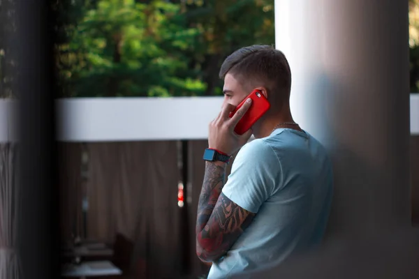 Yakışıklı adam dışarıda cep telefonuyla konuşuyor. Kolunda dövmesi ve akıllı saati olan modern stil sahibi genç adam telefona cevap veriyor. Akıllı telefonu kırmızı bir çantada. Arka plan