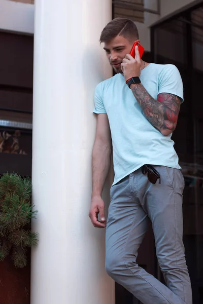 Yakışıklı adam dışarıda cep telefonuyla konuşuyor. Kolunda dövmesi ve akıllı saati olan modern stil sahibi genç adam telefona cevap veriyor. Akıllı telefonu kırmızı bir çantada. Dikey 