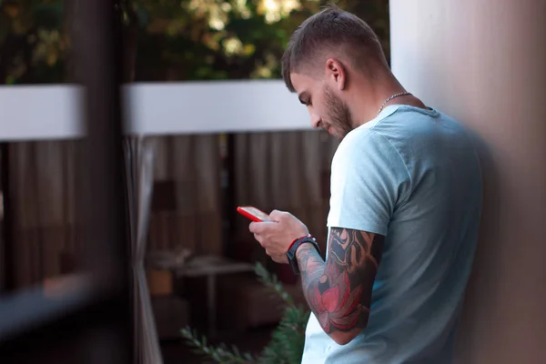 Yakışıklı adam dışarıdaki cep telefonundan mesaj atıyor. Kolunda dövmesi ve akıllı saati olan modern stil sahibi genç adam telefonda bir mesaj yazıyor. Akıllı telefonu kırmızı bir çantada..