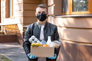 Siyah maskeli ve elleri tıbbi eldivenli genç adam elinde yiyecek dolu karton bir kutu tutuyor. Koronavirüs salgını ve salgın sırasında market alışverişi. Sokakta bağış kutusuyla gönüllü çalışan.