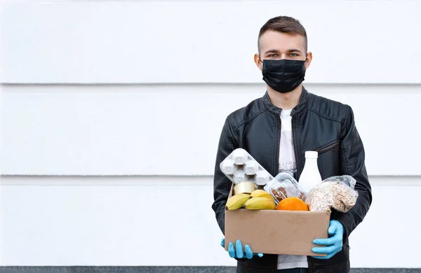 Courier man in face mask with food box in his gloved hands outside. Coronavirus food delivery. Safety contactless delivery. Supplies box with donation food: cereals, eggs, dairy products, and fruits.