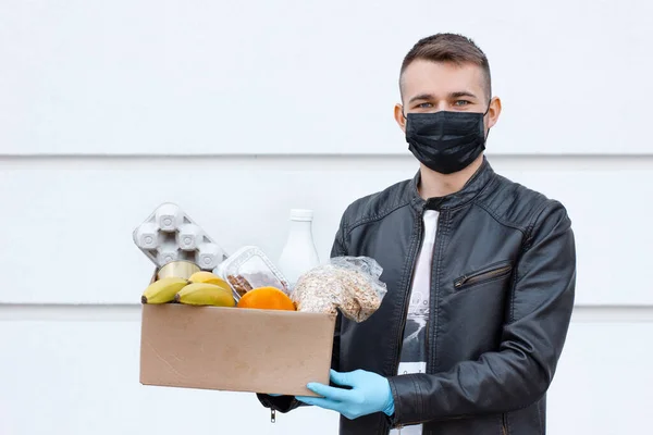 Courier man in face mask with food box in his gloved hands outside. Coronavirus food delivery. Safety contactless delivery. Supplies box with donation food: cereals, eggs, dairy products, and fruits.