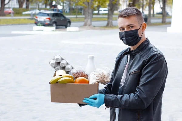 Siyah maskeli ve elleri tıbbi eldivenli genç adam elinde yiyecek dolu karton bir kutu tutuyor. Koronavirüs salgını ve salgın sırasında market alışverişi. Sokakta bağış kutusuyla gönüllü çalışan.