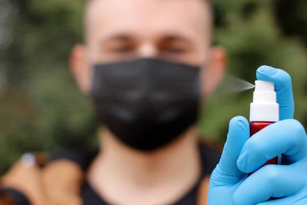 Antiseptic bottle in gloved hand close up. Man in medical gloves and protective face mask spray antiseptic. Personal care product, hand disinfectant. Blurred face in a respiratory mask on background.