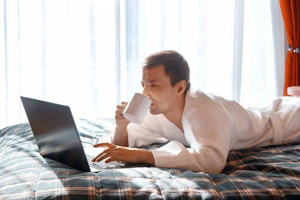 Bornozlu yakışıklı genç adam dizüstü bilgisayarda postaları kontrol ediyor ve evde sabah kahvesini içiyor. Evden çalışıyorum. Mesafe öğrenme. Coronavirus karantinası sırasında iş toplantısı. Evde kal..