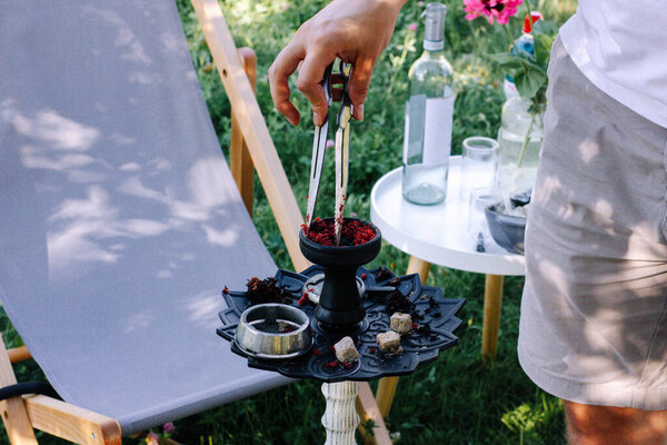 Man's hand distributes red tobacco in a hookah bowl background of summer picnic. Hookah preparation for smoking and fun at summer picnic. Metallic hookah bowl with charcoal inside. 