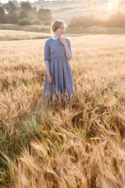 Niña en vestido azul de pie al amanecer en el campo de las orejas en la luz dorada del sol. colina y paisaje de campo en el fondo — Foto de Stock