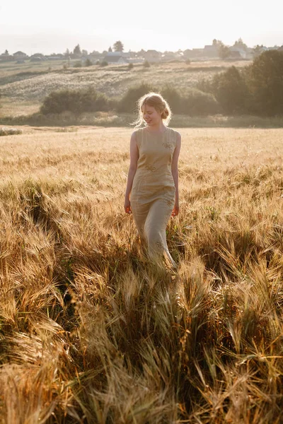 La muchacha joven feliz bonita en el vestido amarillo camina en el campo de las orejas en los rayos del sol naciente. arboleda y pueblo en el fondo — Foto de Stock