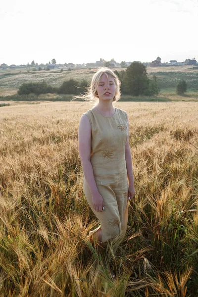 La muchacha joven bonita en el vestido amarillo se encuentra en el campo de las orejas en los rayos del sol naciente. arboleda y pueblo en el fondo — Foto de Stock