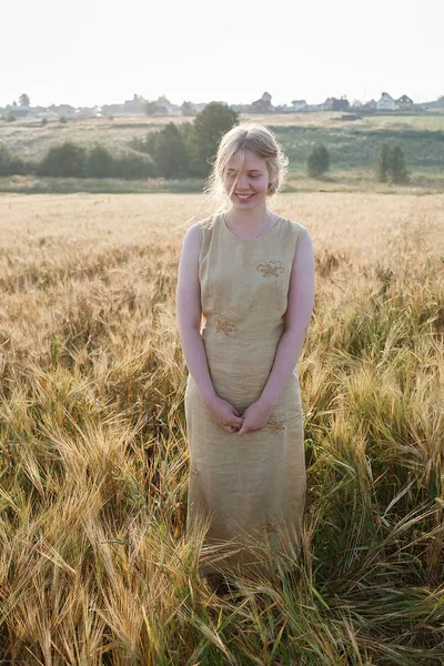La muchacha joven bonita en el vestido amarillo se encuentra en el campo de las orejas en los rayos del sol naciente. arboleda y pueblo en el fondo — Foto de Stock