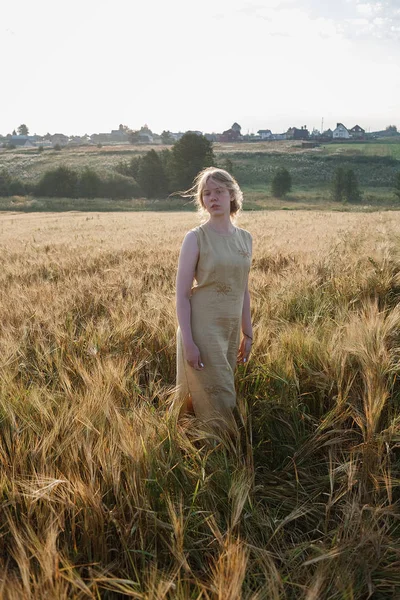 La muchacha joven bonita en el vestido amarillo se encuentra en el campo de las orejas en los rayos del sol naciente. arboleda y pueblo en el fondo — Foto de Stock