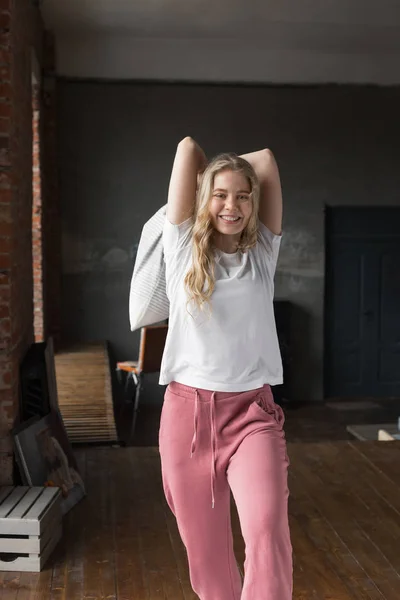 Cute girl ubrana w różowe spodnie piżamy i biały t-shirt na łóżku z poduszką w pobliżu okna, uśmiechnięta i poduszki walki — Zdjęcie stockowe