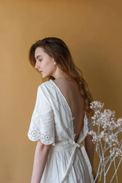 Hermosa joven con el pelo largo en vestido blanco posando sobre fondo beige. Flores blancas en primer plano fuera de foco — Foto de Stock