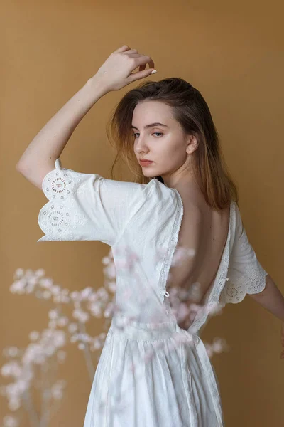 Vacker ung flicka med långt hår i vit klänning poserar på beige bakgrund. Vita blommor på förgrunden ur fokus — Stockfoto
