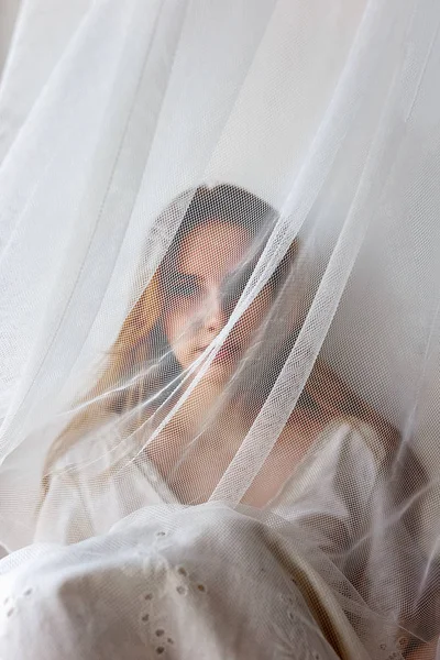 Pretty young sensual girl in white dress hiding behind curtain. Hidden beauty concept