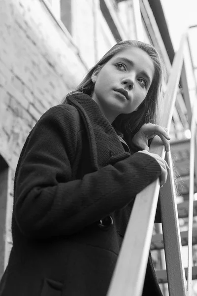 Chica joven con abrigo negro de pie en las escaleras con pared de ladrillo en el fondo. Concepto de soledad. Imagen en blanco y negro —  Fotos de Stock