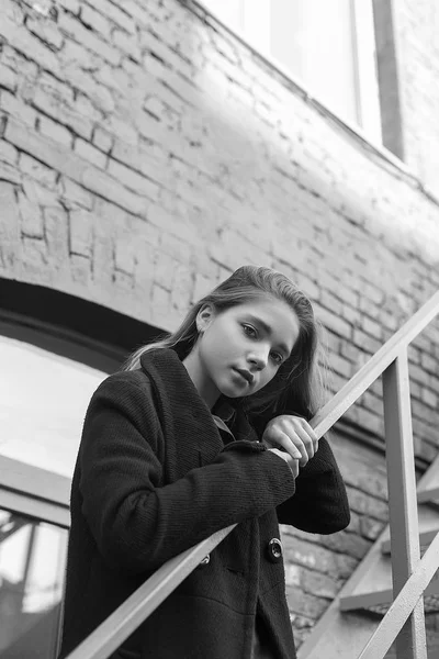 Νεαρό κορίτσι με μαύρο παλτό στέκεται στις σκάλες με τούβλα τοίχο στο παρασκήνιο. Η έννοια της μοναξιάς. Ασπρόμαυρη εικόνα — Φωτογραφία Αρχείου