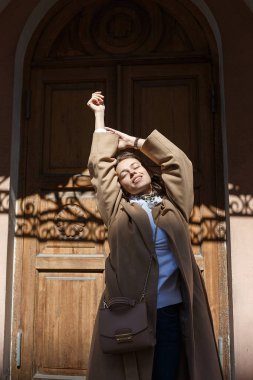 Bir binanın Vintage ahşap kapı önünde kendini germe güneşli bir günde bej ceket genç güzel kız.