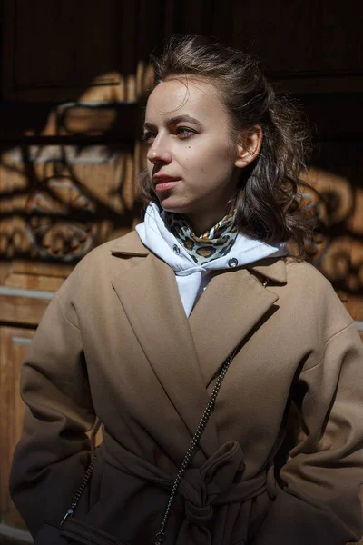 Retrato de estilo de rua de mulher jovem em casaco bege em um dia ensolarado posando na frente de uma porta de madeira vintage de um edifício — Fotografia de Stock