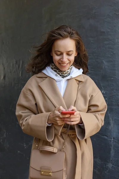Πορτρέτο του δρόμου της νεαρής χαμογελαστή γυναίκα φορώντας μπεζ παλτό χρησιμοποιώντας το έξυπνο τηλέφωνο της σε εξωτερικούς χώρους. Σύγχρονη διαδικτυακή επικοινωνία — Φωτογραφία Αρχείου