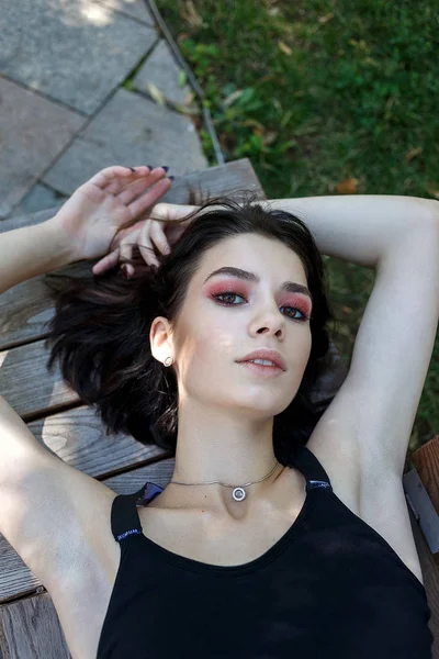 young sexy brunette girl with dark hair, brown eyes, sensual lips, red shadows on her lids in black top lying on bench