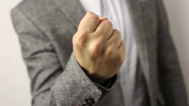 Gri ceketli, beyaz arka planda benekli gömlekli, katı beyaz bir adam yumruğunu kaldırıyor. Kameranın önünde erkek yumruğunun titremesine yakın çekim. Tehdit jesti, saldırganlık konsepti, uyarı, ceza