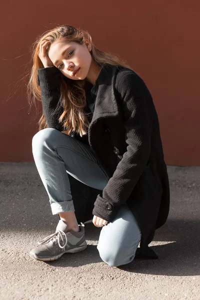 Ragazza in cappotto nero, jeans nascosti sotto e scarpe da ginnastica siede sul pavimento contro un muro rosso e guarda la fotocamera — Foto Stock