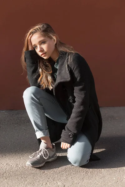 Menina de casaco preto, jeans enfiado abaixo e tênis senta-se no pavimento contra uma parede vermelha. ela parece pensativa e chateada — Fotografia de Stock