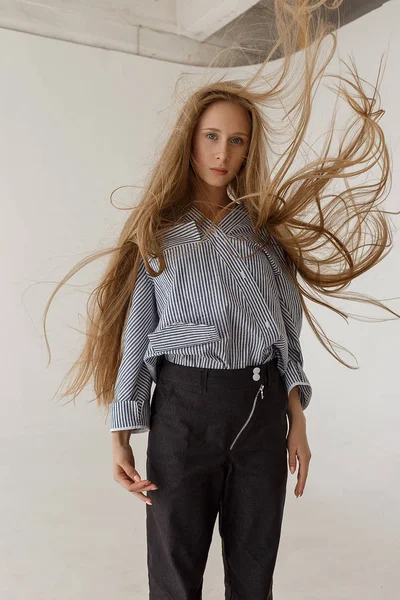 Young teenage girl stands in front of white cyclorama in black jeans and striped oversize shirt with her hair fluttering — Stock Photo, Image
