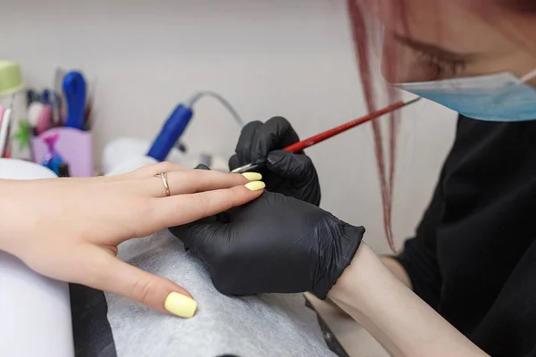 Nail master wearing a mask and black gloves applying brush on acrylic nails in the salon. Closeup