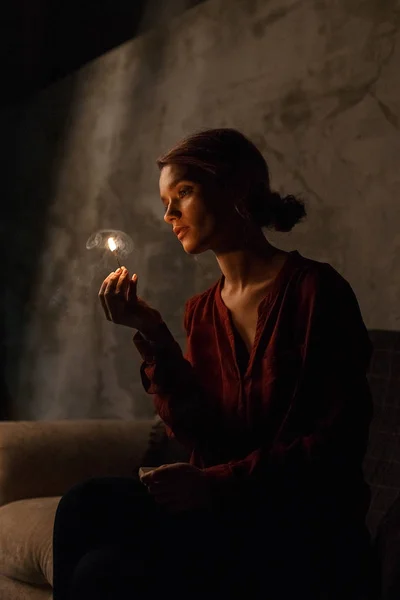 Pretty young girl in red shirt sits in dark gloomy room, looks thoughtfully at fire of burning match and holds matchbox