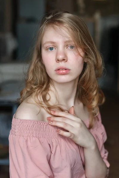 Menina loira atraente nova com olhos azuis em vestido rosa na oficina, segurando a mão no peito e olhando para a câmera — Fotografia de Stock