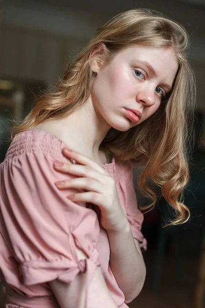 Joven chica rubia bonita con ojos azules en vestido rosa, cabeza inclinada, sosteniendo la mano en el pecho y mirando a la cámara — Foto de Stock