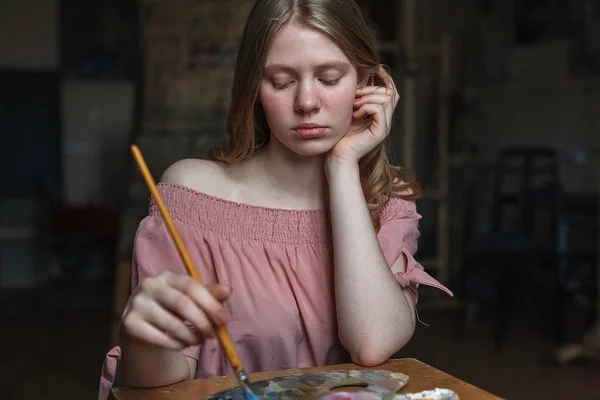 Junges hübsches blondes Mädchen in rosa Kleid legt Kopf auf den Arm und mischt Farben mit Pinsel auf Palette im Kunstatelier — Stockfoto