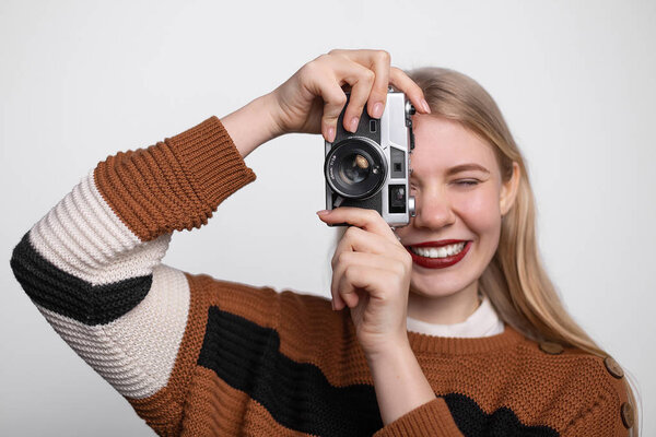 Young pretty blonde girl with bright red lips smiling, holding vintage photo camera, taking picture on white background
